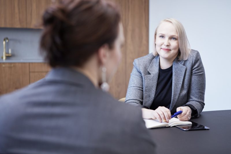 Henkilöt istuvat vastakkain. Toinen henkilö hymyilee.