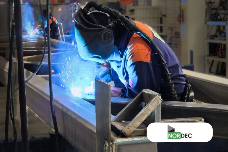 Kuvassa on suojavarusteisiin ja hengityssuojaimeen pukeutunut hitsaaja hitsaamassa teräspalkkia